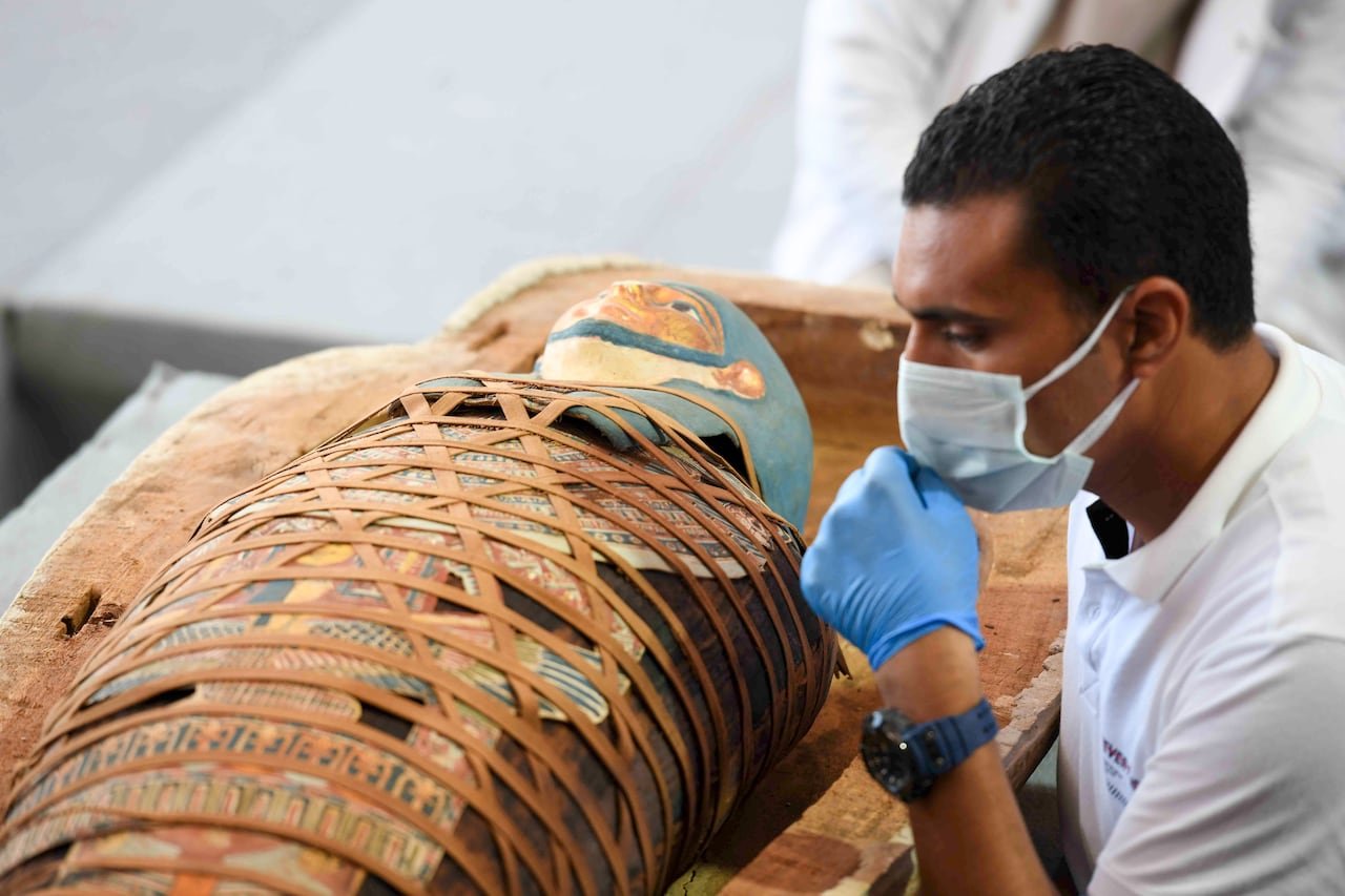 default-1629 An archaeologist inspects an Egyptian mummy that is wrapped in an elaborate and colourful burial shroud. The archaeologist is wearing a medical mask and gloves.