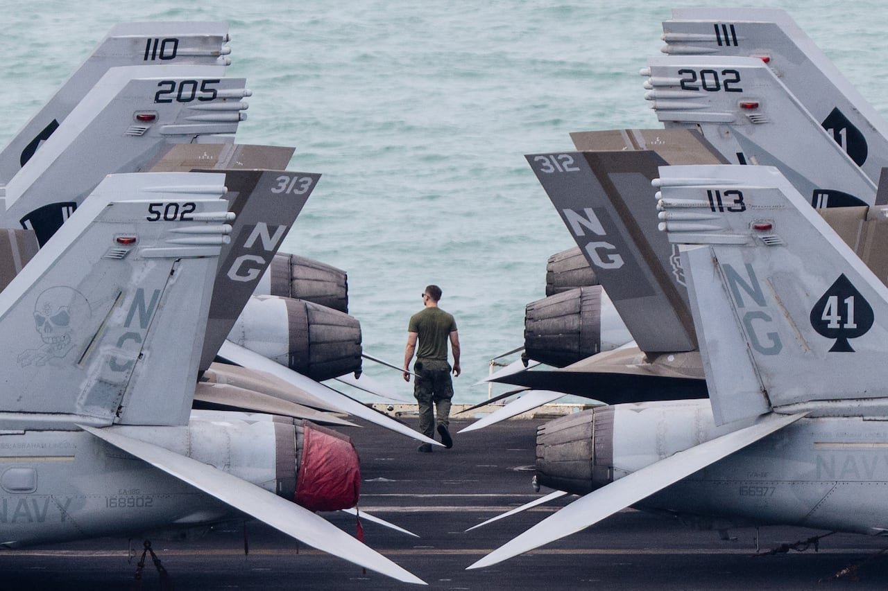 A sailor in green walks toward the water aboard a ship, framed on either side by the numbered tails of grey navy jets.